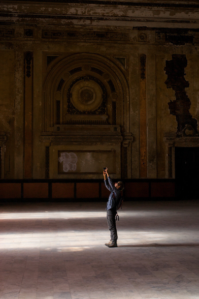 A woman stretches to take a picture inside an abandoned station hall. Light spills from the windows onto the floor, otherwise the interior is dark