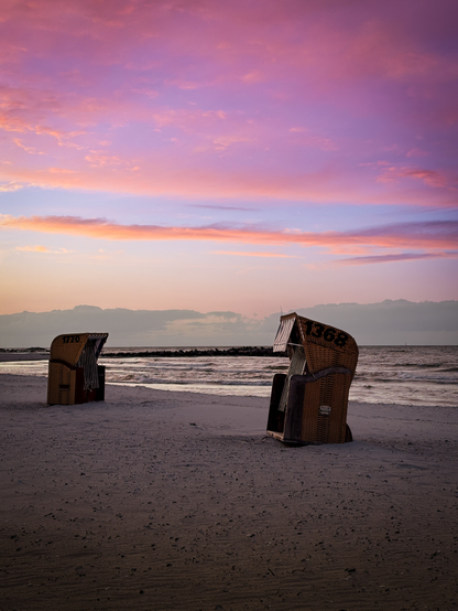 Zwei Strandkörbe stehen bei Sonnenuntergang am feinen hellen Sandstrand. Die Sonne ist hinter einem dichten Wolkenband nicht zu sehen, aber ihr Licht strahlt den Himmel darüber orange anX weiter oben scheinen rosa Wolken. Ein Strandkorb steht hinten links eher Hintergrund, er ist aufs Meer gerichtet. Vorne rechts steht ein Strandkorb, der vom Meer abgewandt ist.