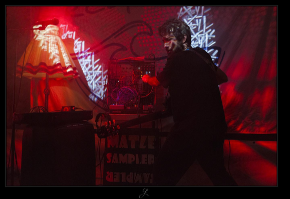 On a red-lit stage, a bearded man in dark clothing can be seen from behind at an angle. He has an electric guitar slung over his shoulder and is working on controls and switches on a rack equipped with numerous electronic devices with LEDs, plugs, and cables. Below the rack, there is a dark poster with bright, bold letters that says "MATZE SAMPLER". To the left of the rack, a mixing console stands on a suitcase. In the background behind the suitcase is a large, old-fashioned floor lamp with a lampshade. Red and white patterns from moving stage lights can be seen on the wall. The colors are mainly red and black.

AI disclaimer: Using my work, its meta data, written or derived description to create media with or train AI based systems is prohibited.