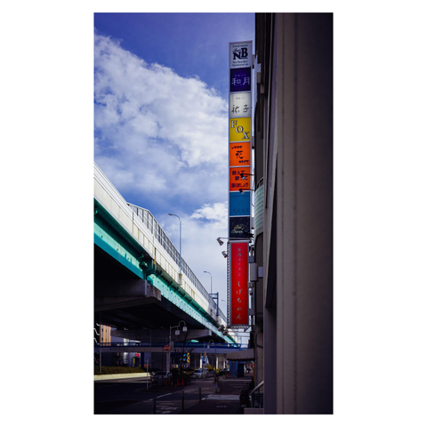 A color photo of a Nagoya street scene with a tall vertical signpost attached to a building. The signs are stacked in different colors, including bright orange, deep red, and yellow, standing out against a clear blue sky. On the left side of the frame, an elevated highway in teal stretches into the distance. The composition brings together industrial structure and seasonal color, suggesting autumn through signage rather than trees.