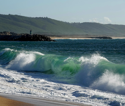 Das Bild zeigt eine beeindruckende Küstenlandschaft mit einem großen, kräftigen Wellenbruch, der sich an den Strand stürzt. Die Wellen sind türkisgrün und mit Schaumkronen bedeckt, die sich in der Luft verteilen. Im Hintergrund erhebt sich ein grünes Hügelgebirge, das sanft in die Ferne übergeht. Links im Bild ist ein Leuchtturm auf einem Steindamm zu sehen, der als Schutz vor den Wellen dient. Der Himmel ist klar und blau, was auf einen sonnigen Tag hinweist. Die Szene vermittelt ein Gefühl von Naturgewalt und Schönheit