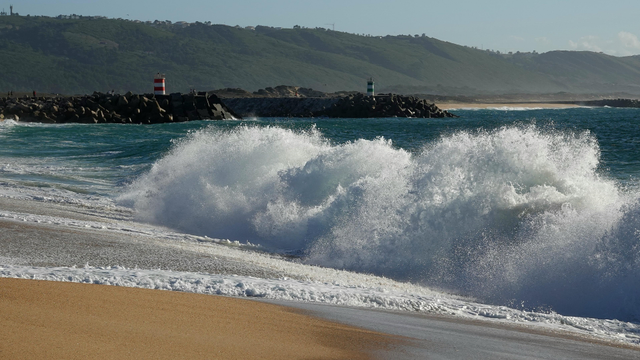 Das Bild zeigt eine beeindruckende Küstenlandschaft mit einem breiten, goldenen Sandstrand im Vordergrund. Die Wellen des türkisblauen Meeres brechen mit großer Kraft an den Strand, erzeugen spritzendes Weiß und Schaum. Im Hintergrund ist ein Steinwall zu sehen, der das Ufer schützt, und auf ihm stehen zwei Leuchttürme, einer rot-weiß gestreift und der andere weiß-grün. Die Leuchttürme dienen als Orientierungspunkte für Schiffe. Dahinter erstrecken sich sanfte Hügel mit grüner Vegetation, die die Küste säumen. Der Himmel ist klar und blau, was auf einen sonnigen Tag hinweist.