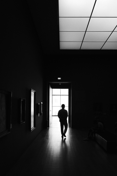 A black and white, slightly low-angle shot of a hallway. 

The hallway is dark, with the walls and floor a deep black, emphasizing the contrast with a sliver of light coming from an open doorway.  

A person is walking away from the camera towards the open door and bright light on the other side. The figure is a silhouette, with no visible details.  

Framed paintings or artwork are affixed along the walls.

The ceiling is a patterned grid of light gray squares. The light from the doorway on the other side of the hallway seems to illuminate the floor creating a bright trail fading back into the dark.