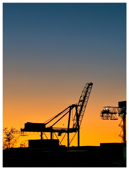 Photo of a shipping crane at sunset, standing in silhouette with its boom raised like a giraffe. The clear sky displays a gradient of color, from a soft blue higher up to a golden yellow closer to the horizon. The crane is flanked by other silhouettes: a tree at left, and a portion of another crane at right, with some illuminated safety lights. Through the middle portion of the crane we can see the tiny figure of a distant helicopter against the sunset. Stacked shipping containers stand in shadow in the foreground.