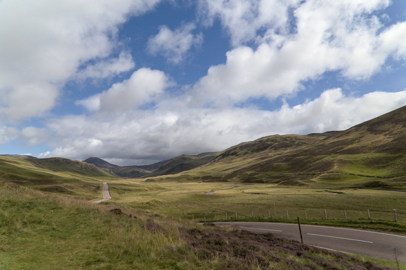 Landscape in the Scottish Highlands. We can see a road going across the mellow mountains of the Highlands, covered with green grass, the sun lighting some part of it through the clouds, quite numerous but letting some vivid patches of blue sky visible