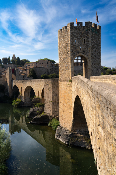Color photo of a medieval stone bridge with a crenellated tower gateway and rounded Romanesque arches, leading over a river to a town beyond. 
