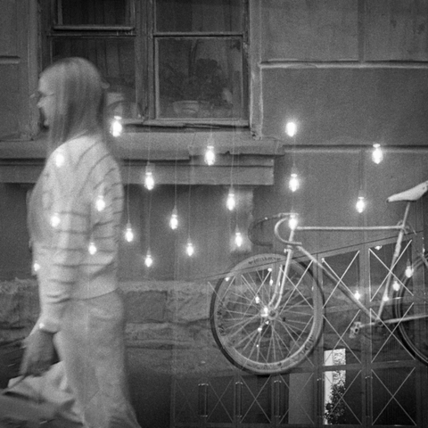 The black-and-white photo shows part of a house wall with a window. A bicycle is parked against the wall. A woman walks past in the foreground. Superimposed on the photo is another photo of a cafe interior with vintage light bulbs glowing.