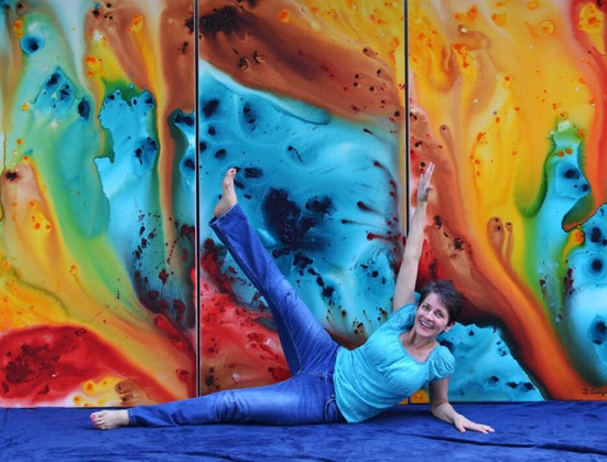 A female on the ground in a joyful position in front of a colorful abstract painting.