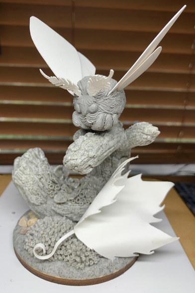 Photo of a sculpture of a chonky moth with large antennae perched on a curved branch covered in lichen with a curled up lead in the foreground. It’s unpainted, showing the gray clay and white elements which are actually paper. 