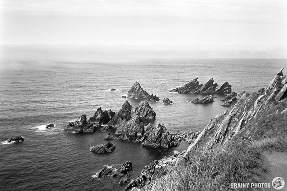 A black-and-white film photo of the coastline - a series of jagged rocks protrude from the water.