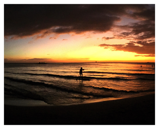 Photo on a beach at sunset, looking out to sea. The orb of the sun isn’t visible but its golden glow fills the sky, shining on the water and backlighting a mass of low clouds. The waterline curves along the foreground against a black field of shadowed sand. At the center of the image, a paddle boarder stands up in the sea, silhouetted against golden waves, pushing an oar into the water. At right are tiny dots of distant swimmers. A ship with a dot of green light sits at the horizon.