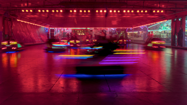 Foto einer Autoscooter-Bahn im Betrieb. Die Lichter leuchten rot-orange, im Vordergrund fährt ein einzelner Scooter von rechts nach links und ist durch die lange Belichtungszeit nur als bunter Schemen zu erkennen.