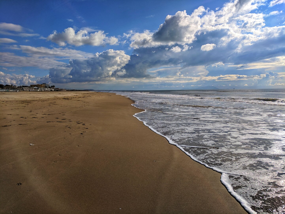 A serene, wide-angle photograph of a sandy beach meeting the ocean under a dramatic sky. The golden sand stretches along the shoreline, gently curving toward the calm, rolling waves of the sea. Above, the sky is filled with a mix of fluffy white clouds and darker, billowing storm clouds, creating a striking contrast. In the distance, a few buildings and structures are visible along the coastline, adding a touch of human presence to the otherwise natural landscape. The scene captures the tranquillity and vastness of the beach, with the interplay of light and shadow enhancing its beauty.