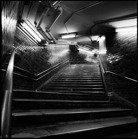 Black and white photograph of a station staircase, taken from the bottom of the flight, with multiple changes of direction, and very dramatic lighting.