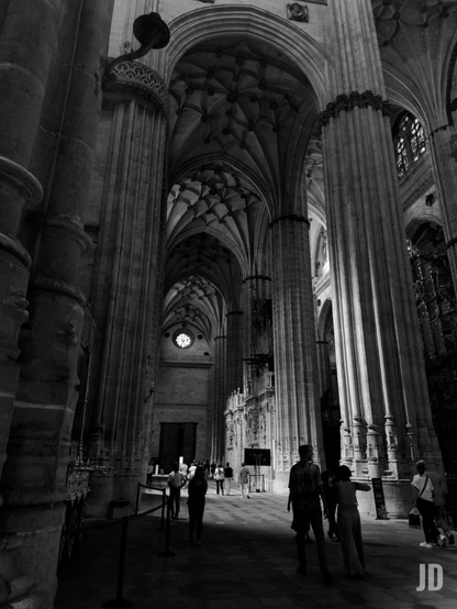 La imagen en blanco y negro muestra el interior majestuoso de una gran catedral o iglesia gótica, vista desde un ángulo bajo que enfatiza su altura y profundidad. Un pasillo largo y oscuro se extiende hacia el fondo, flanqueado por imponentes columnas estriadas que se elevan hacia arcos ojivales. El techo está adornado con una intrincada bóveda de crucería estrellada. La iluminación es dramática, con el primer plano y los laterales en sombra, mientras que la luz se filtra desde ventanas altas y un rosetón circular visible al final del pasillo. A la derecha, se observa una elaborada estructura lateral con ornamentación detallada. Varias personas, pequeñas en comparación con la escala del edificio, caminan o se detienen en el pasillo, proporcionando una sensación de escala al monumental espacio.