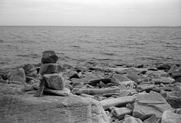 The photograph depicts a rocky shore, with a small artificial stone pyramid visible in the foreground. A lake can be seen in the distance. The sky is overcast, creating an atmosphere of calm and tranquility.