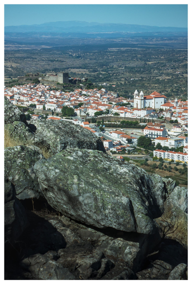 The image depicts a picturesque town with a panoramic view. In the foreground, large, rugged rocks with patches of green moss and lichen are visible, adding texture to the scene. The town is characterized by white buildings with red-tiled roofs, densely packed together, creating a harmonious and traditional appearance. A prominent church with two white spires stands out, suggesting its significance in the town's architecture. In the background, a castle-like structure with a tower is visible, indicating historical significance. The landscape extends into the distance, with rolling hills and a clear blue sky, providing a serene backdrop. The overall scene is a blend of natural and man-made elements, showcasing the town's charm and historical context.

Provided by @altbot, generated privately and locally using Ovis2-8B