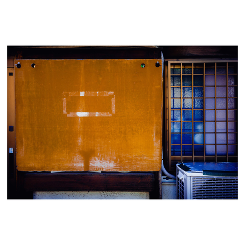 A color photo showing a rectangular orange board hanging on hooks, covering part of a wall. The surface is worn, and a rectangular outline of tape residue suggests something—a label or sign—was once affixed there but is now gone. To the right is a gridded window with blue glass, and below it sits an air conditioning unit.