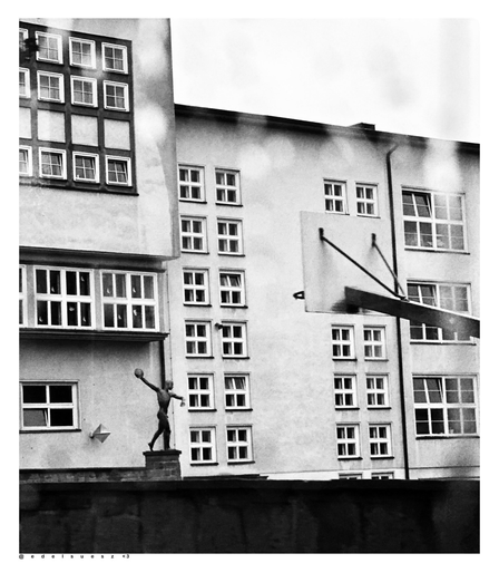 Schwarzweißfotografie: ein helles kastenförmiges Gebäude mit vielen kleinen quadratischen Fenstern, eine Schule aus den 1930er Jahren im Bauhausstil, unterhalb entfernt eine Statue eines Diskuswerfers, rechts daneben im Innenhof ein Basketballkorb mit Halterung von hinten, davor eine dunkle Mauer 