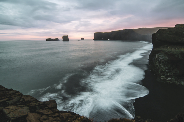 Dramatic view of the Icelandic coastline at sunset, with dark cliffs, black sand beach, and foamy waves rolling ashore. Offshore sea stacks rise from the calm water under a cloudy pink and purple sky.