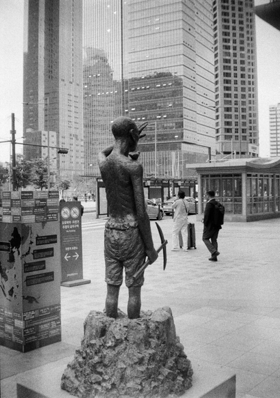 뒤편에서 바라본 강제징용 노동자상. 오른손에는 곡괭이를 들고 있다. 노동자의 무릎 아래는 돌에 파묻혀있는 형상이다. 노동자가 바라보는 방향에는 마천루가 가득 들어찼다.