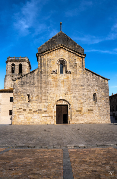 Color photo of the facade of an old Romanesque church with a square tower in the background, facing an empty plaza.