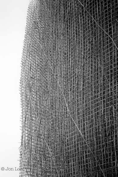 This is a simple abstract black and white photo of a large framework of bamboo scaffolding. 
Covering the entire image is a lattice of vertical and horizontal poles of bamboo scaffolding. There are sixty-five lines of horizontal poles and seven rows of vertical poles, each separated by about one metre. Each line is make of multiple poles each of about three metres long, lashed together with short lengths of binding straps, the sort used to strap a large parcel. There are also numerous diagonal poles lashed to the horizontal and vertical poles for added strength. The scaffold is not two dimensional, there are numerous similar lattices behind at least three and probably more, again to add strength. The entire structure was vast with the height being of greater than thirty metres.
The location: The Hong Kong Convention and Exhibition Centre in Wan Chai (2014).

