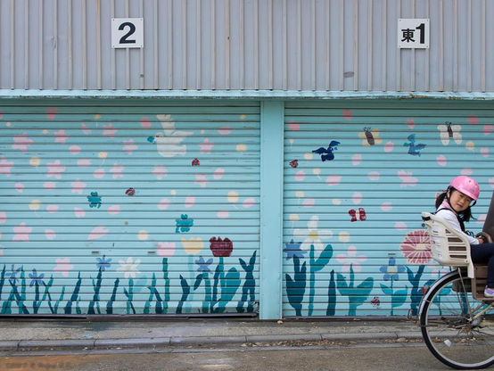 A photo of storage garage doors painted with flowers and butterflies as the background small girl on the back of mama's bicycle about to exit the frame.