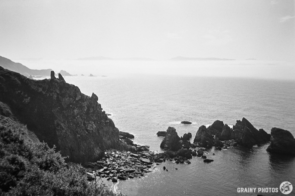 A black-and-white film photo of the coastline - a series of jagged rocks protrude from the water.