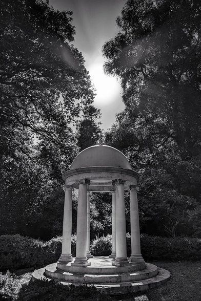 A striking black and white photo of a circular, classical-style stone temple or gazebo, featuring a domed roof supported by six columns. The structure is set on a low, tiered base in a garden or wooded area. Tall, dense trees frame the structure on both sides, with the bright sun directly behind the dome at the top centre, creating a strong lens flare and dramatic contrast.