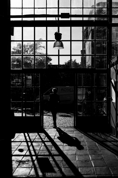 A black-and-white photograph shows a silhouetted figure standing in an open doorway, framed by a grid of glass panes. The high-contrast scene plays with light and shadow, casting the person's shadow across a tiled floor. A large lamp hangs above, faintly lighting the interior. Outside, a cobblestone street, and distant trees suggest a sunny day. The composition is balanced and dramatic, evoking a sense of transition or introspection.