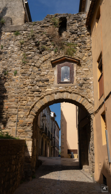 Color photo of an arched opening in a wall through which passes a cobblestone street.  Above the arch is a glass-covered niche holding a small religious figure.    