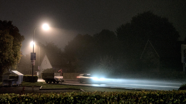 A foggy evening shot, showing a street with a lantern and a car passing by. The car headlights beam is cutting through the fog.