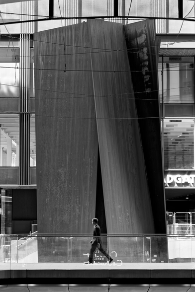 Black and white photograph of a man in a suit walking along a glass walkway in front of a monumental steel sculpture and modern building facade, creating a striking contrast between human scale and architectural mass.