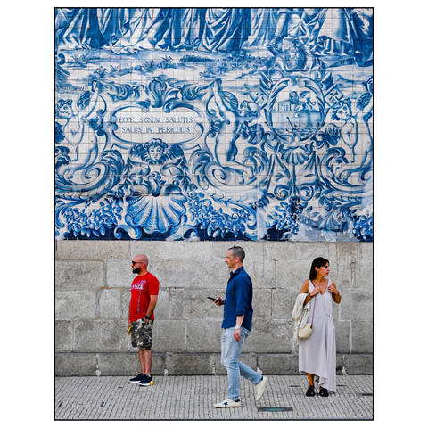 This image shows three people standing in front of a large, decorative tile mural. The mural features traditional Portuguese azulejos—blue and white ceramic tiles—depicting intricate, historical or mythological scenes with elaborate patterns, including figures, sea creatures, and ornate designs. The Latin phrase "ECCE SIGNUM SALUTIS SALUS IN PERICULIS" is prominently displayed on a banner within the mural, which translates to "Behold the sign of salvation, safety in danger."
The three individuals appear to be not noticing each other at all. One is standing looking to the left, the other is walking by watching his smartphone, the third is standing on the right, looking to the right. (draft by mistral.ai, edited by author)