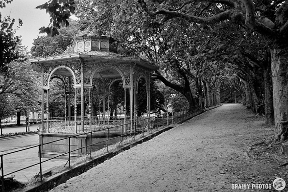 A black-and-white film photo of a path in a wooded section of the park. There is an ornate bandstand on the LHS