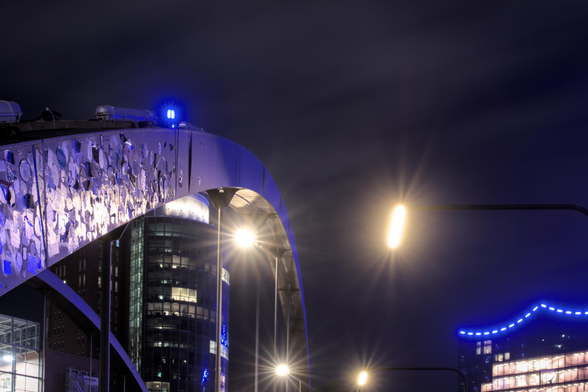 Bogenförmige Brückenkonstruktion mit reflektierenden Platten und blauem Licht vor der beleuchteten Elbphilharmonie in der HafenCity bei Nacht