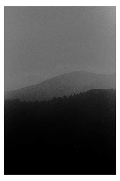 A grainy black and white photograph of distant mountain ridges fading into mist. The dark silhouette of the foreground hills contrasts with the lighter tones of the background, evoking a quiet, contemplative mood. A minimal landscape for #SilenceSunday.