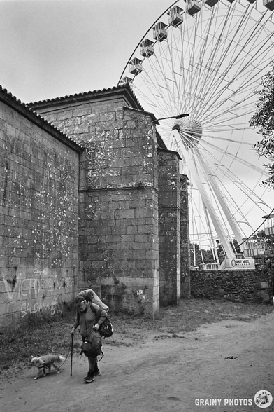 A black-and-white film photo of an older pilgrim with his dog walking past a church in Santiago