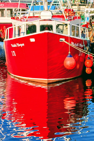 A close-up, vertical shot of the bright red bow of the fishing trawler docked in a harbour. The boat's vibrant red hull is intensely reflected in the calm, deep blue water below, creating a striking contrast and a shimmering, inverted image. Two small, bright orange buoys hang from the boat's side, mirrored in the reflection.