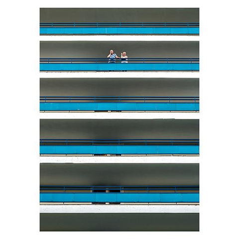A man in a blue and white striped shirt stands on the 4th floor of a modern, five-story hotel building, next to him a woman in a white shirt. The railing of the hotel balcony is blue, and the floors of the balconies are white. This makes the building look like a blue and white striped thing from below. The couple is wearing clothes that match the color of the hotel building exactly.