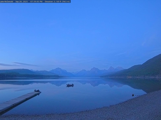 The view this evening of Lake McDonald in Glacier National Park is blue and symmetrical. The lake surface is mirror-like, reflecting mountains and clouds above. On the left, park visitors soak in the scene from the dock. A boat anchors the center of the image and is itself reflected by the water. 