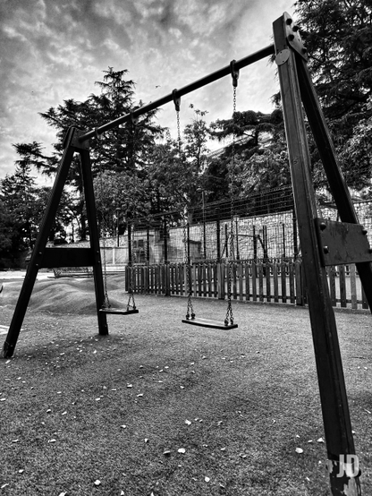 Fotografía en blanco y negro de un parque infantil, tomada desde un ángulo bajo. En primer plano, una estructura de columpios metálica y oscura sostiene dos asientos vacíos, que cuelgan de cadenas. El suelo del parque es de goma o césped artificial, salpicado de hojas dispersas. Al fondo, se distingue una valla de madera y otra de malla metálica, detrás de las cuales se ven árboles frondosos y un edificio. El cielo está cubierto de nubes densas, lo que contribuye a una atmósfera tranquila y ligeramente melancólica en este parque aparentemente desierto."