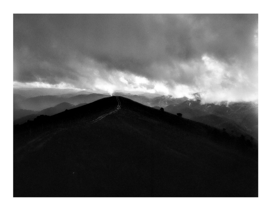 A dramatic black-and-white photograph of a mountain ridge with a narrow path leading into distant storm clouds, taken from a high vantage point with layered hills fading into the mist.