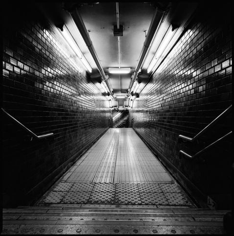 black and white moody picture of a staircase in a tokyo metro station. the lighting is dramatic, bright white reflecting off dark tiled walls. 