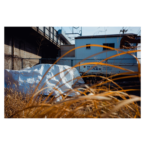 A color photo of an industrial scene near train tracks in Nagoya. The foreground is filled with tall, dry golden-brown grass in sharp focus. Behind it, partly obscured, is a truck with Japanese text on the side and a large white tarp covering an unidentified object. A chain-link fence separates the lot from the railway structure and a simple white building.