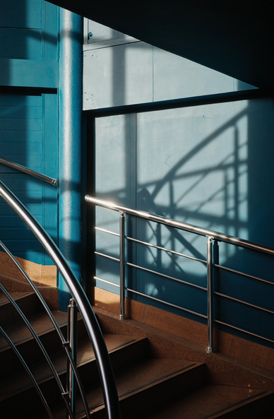 The photograph depicts a staircase with metal railings leading upward. The walls are painted blue, and the steps are made of brown material. Light coming through the window creates interesting shadows on the walls and railings, adding depth and volume to the image.