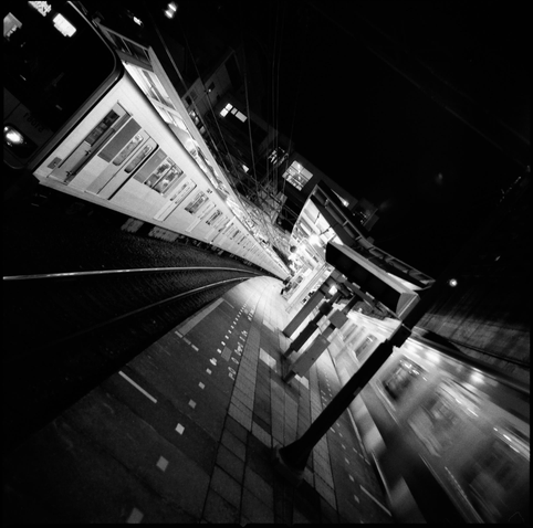 black and white photographs of two tokyo metro trains, the picture is tilted at an angle of roughly 45º. the train o the left side is leaving the station and blurred by motion. the platform curved into the centre of the frame. it is night... and for once it hasn't been raining.
