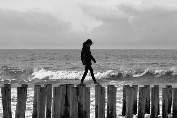 das bild zeigt eine schwarz-weiß-aufnahme eines kindes mit langen haaren, das barfuß auf einer reihe von holzpflöcken am strand balanciert. im hintergrund ist das meer mit leichten wellen und einem bewölkten himmel zu sehen. 
ob der bewölkung ist das bild verhältnismäßig stark kontrastiert. das kind ist fast silhouettenhaft dunkel vor dem hintergrund. die pflöcke befinden sich im vordergrund und ragen aus dem wasser, das zwischen ihnen hindurchfließt.