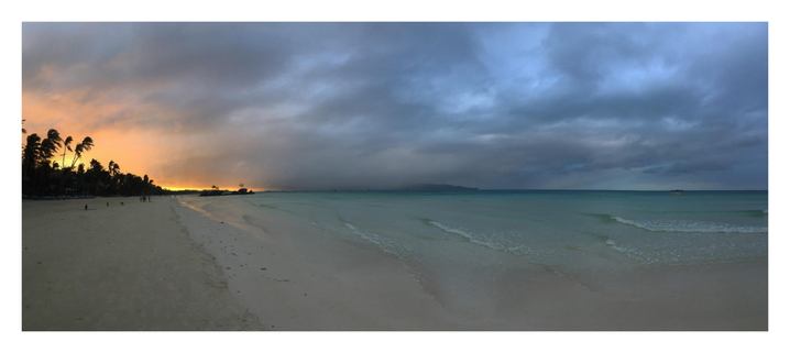 Panorama photo of an island beach at sunrise. The shore of fine white sand is at left, the ocean at right. The beach has a very gradual slope allowing it to stretch far into shallow water, which rolls to shore in lines of gentle waves. The sun peers through palm trees at left frame, silhouetting them in gold against an overcast sky of low blue clouds. In the distance are tiny figures of people strolling on the sand and stray dogs sniffing for food. A line of volcanic rocks sits in the water close to shore, topped by a few trees. At right, a fishing boat sits close to the horizon.